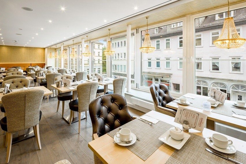 Helles Hotelrestaurant mit gepolsterten Stühlen, großen Fenstern mit Blick auf Stadtgebäude und eleganten Kronleuchtern an der Decke