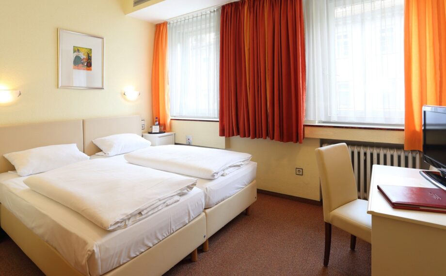 Helles Hotelzimmer mit zwei Einzelbetten, weißer Bettwäsche, orangen Vorhängen, Schreibtisch mit Stuhl und Wandlampen zum Lesen.