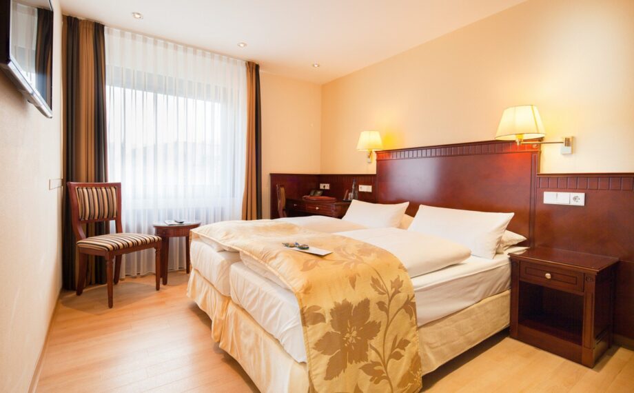 Helles Hotelzimmer mit zwei Einzelbetten, elegantem Holz-Kopfteil, gestreiftem Stuhl und durchsichtigen Vorhängen für Tageslicht