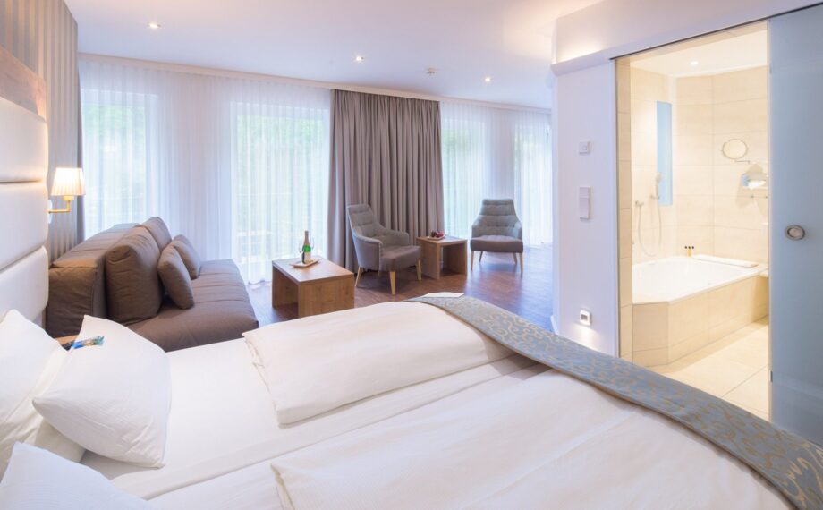 Helle Hotelsuite mit bequemem Doppelbett, Sitzbereich mit Sofa und Sesseln sowie modernem Badezimmer mit Badewanne.