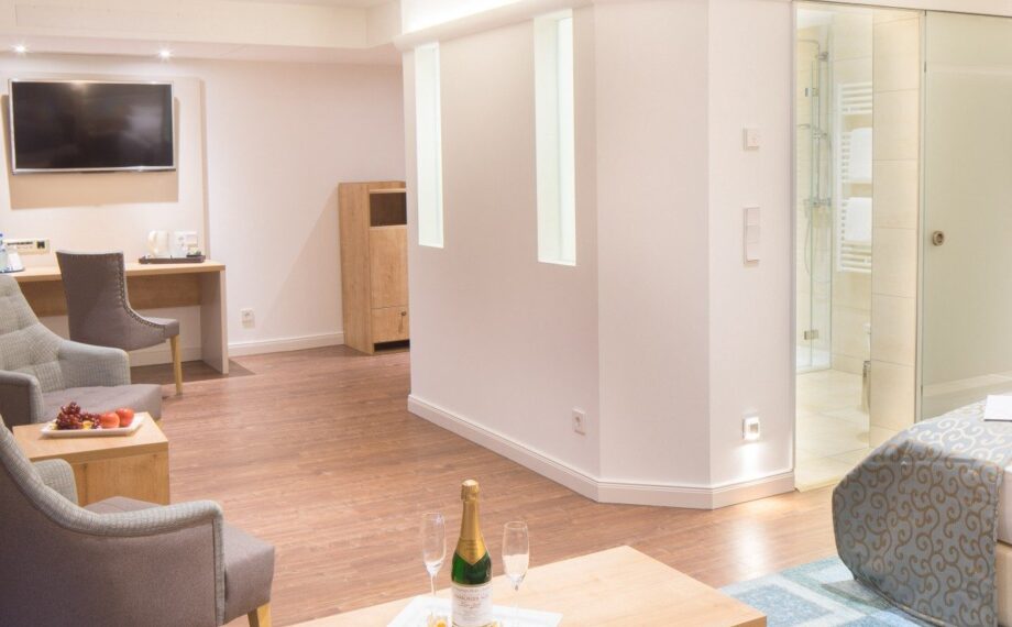Moderne Hotelsuite mit Holzfußboden, Sitzbereich, Wand-TV, gemütlichem Bett und einem Tablett mit Champagner auf dem Couchtisch