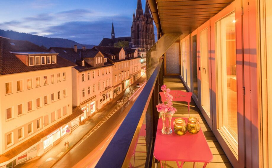 Gemütlicher Balkon mit pinkem Tisch und Stühlen, Kerzen und Blumen mit Blick auf historische Stadt und Kirchtürme bei Dämmerung