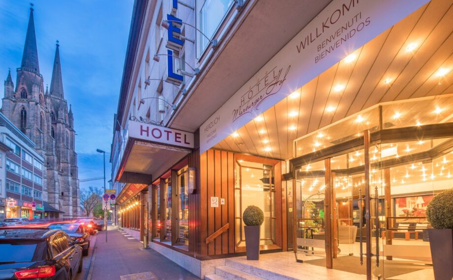 Einladender Hoteleingang mit leuchtender Beleuchtung, Glastüren und Blick auf eine Stadtstraße nahe einem historischen Kirchturm bei Dämmerung