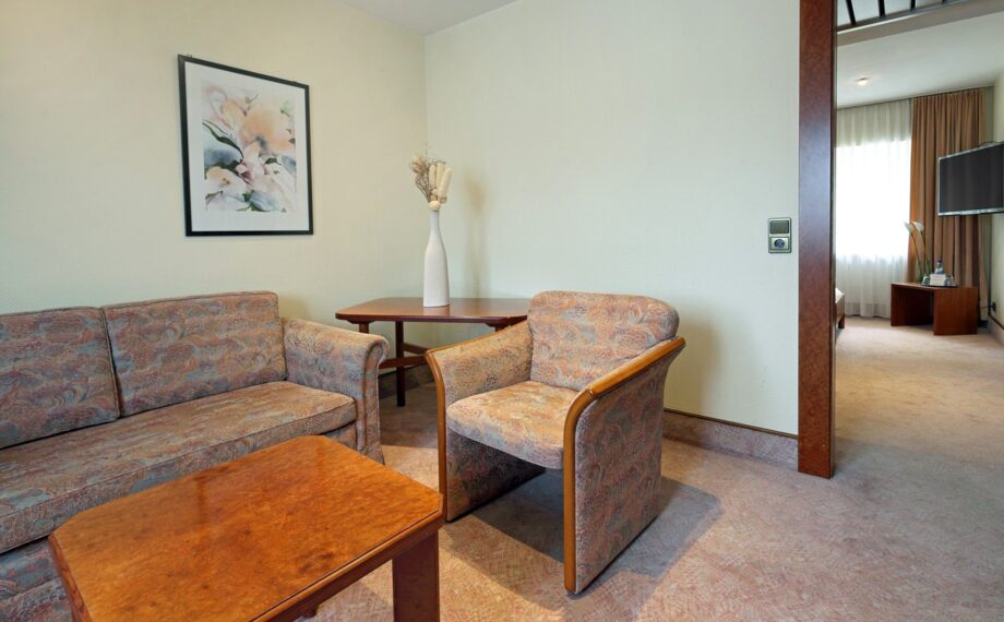 Wohnbereich einer Hotelsuite mit gemustertem Sofa und Sessel, Holztisch, Beistelltisch mit Vase und Tageslicht aus angrenzendem Zimmer