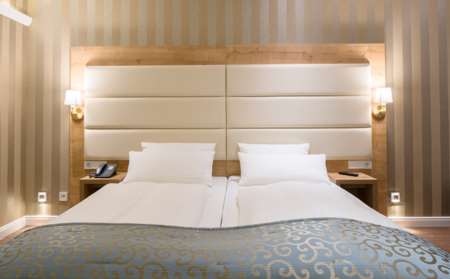 Modernes Hotelzimmer mit zwei Einzelbetten, gepolstertem weißem Kopfteil, beige gestreifter Tapete, Nachttischlampen und blauem Bettüberwurf mit Muster