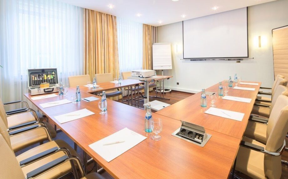 Heller Konferenzraum mit U-förmigen Holztischen, Ledersesseln, Wasserflaschen, Beamer und viel Tageslicht durch große Fenster
