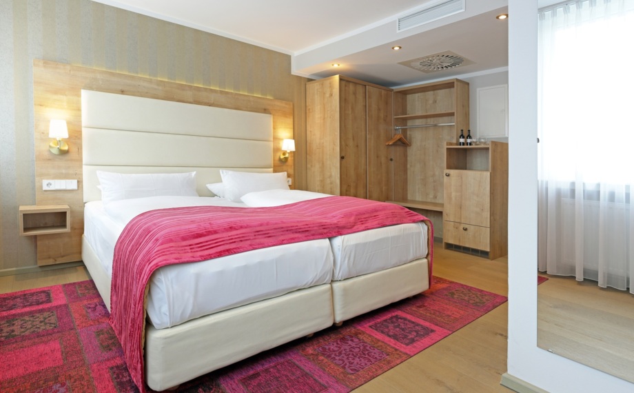 Modernes Hotelzimmer mit großem Doppelbett, weicher Kopfteil, rosa Überwurf und Teppich, Holzkleiderschrank und Tageslicht am Fenster