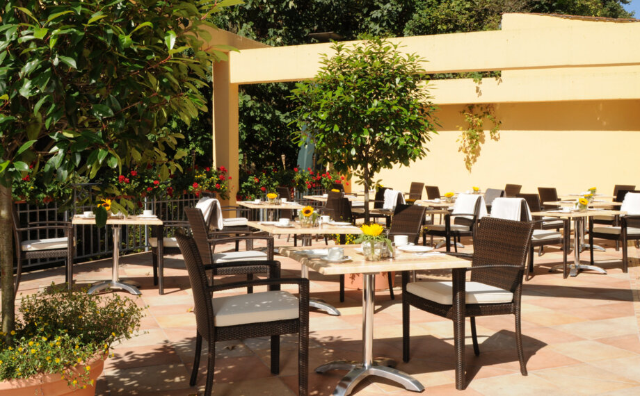 Sonnige Außenterrasse des Hotels mit Korbsesseln, Marmortischen, Topfpflanzen und kleinen gelben Blumenarrangements
