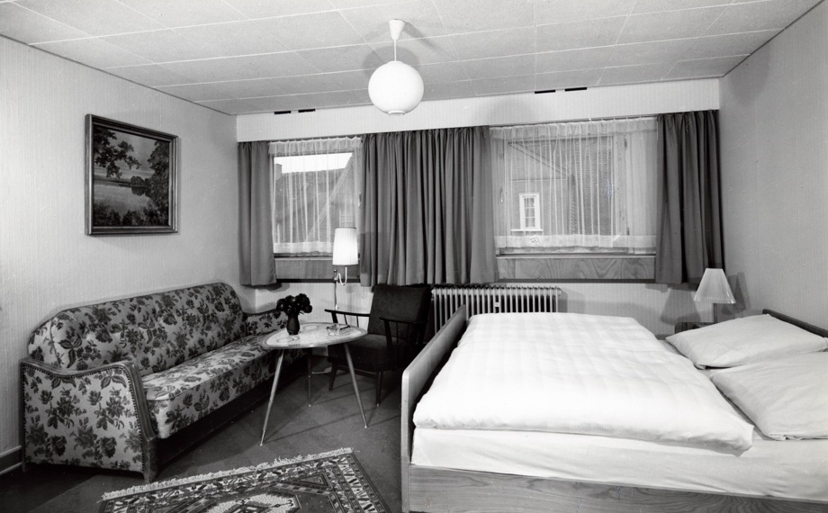 Gemütliches Hotelzimmer mit Doppelbett, floraler Couch, Sessel, kleinem Tisch mit Blumen, Gardinen, Heizung und Wandbild