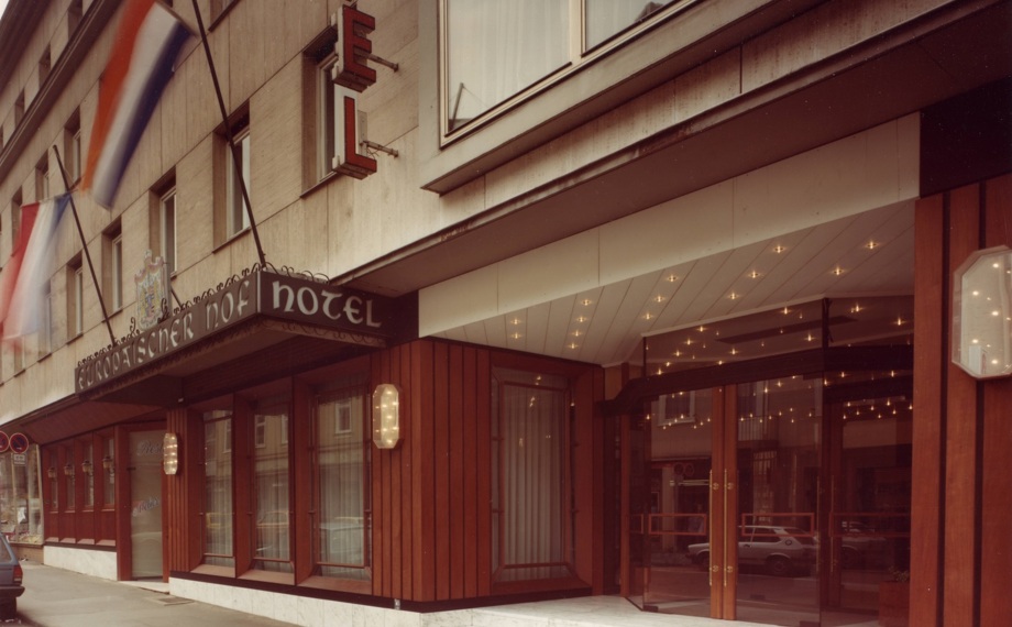 Klassischer Hoteleingang mit polierten Holzverkleidungen, Leuchtreklame, Marmorstufen und sanfter Deckenbeleuchtung an einer Stadtstraße.