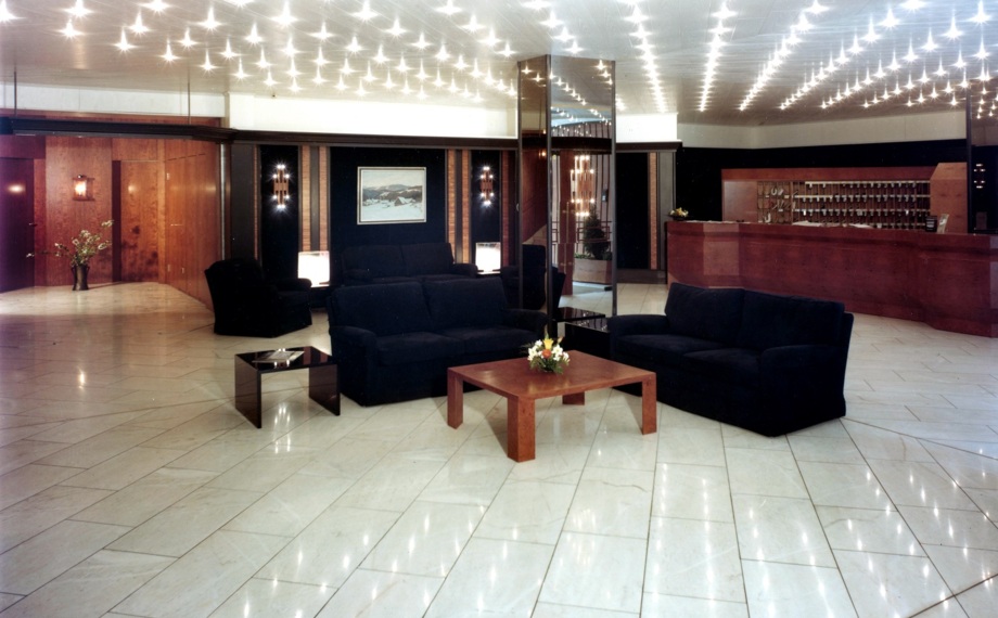 Großzügige Hotellobby mit glänzendem Fliesenboden, dunklen Sofas um Holztische, eleganten verspiegelten Säulen und warm beleuchtetem Empfangstresen