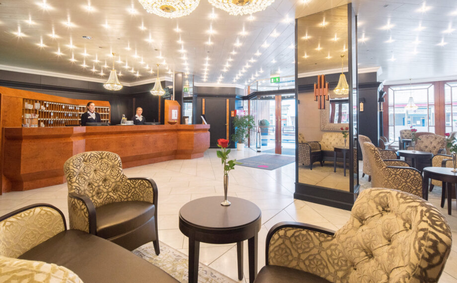 Heller Hotellobby mit poliertem Holzeingangstresen, freundlichem Personal, eleganten Sesseln, Kronleuchtern und frischen Rosen auf den Tischen