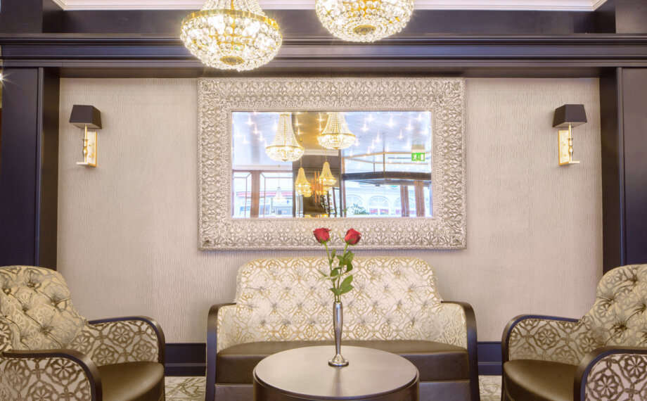 Elegante Hotellounge mit gemusterten Sesseln, einer Couch, rundem Tisch mit silberner Vase und zwei roten Rosen sowie verziertem Spiegel an der Wand