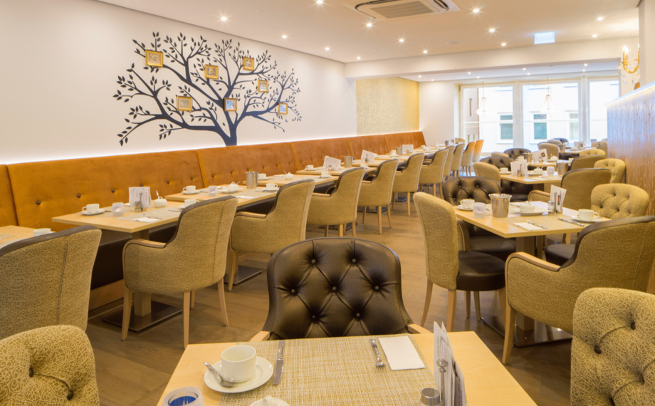 Heller Hoteldiningbereich mit gesteppter Polsterung, senffarbenem Bankett, Wanddekor in Form eines Baums und gedeckten Frühstückstischen.