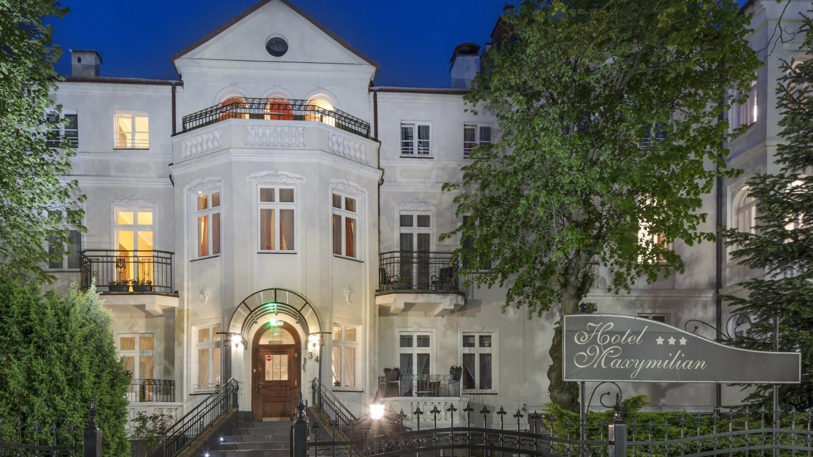 Elegancka zabytkowa willa butikowego hotelu nocą, podświetlane wejście, kuta brama, schody, ogród i balkony — Hotel Maxymilian