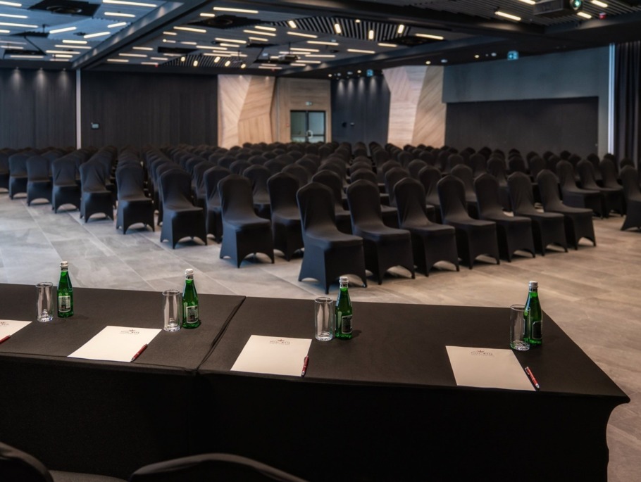Nowoczesna sala konferencyjna z rzędami krzeseł w czarnych pokrowcach, frontowy stół z wodą, szklankami i notesami.