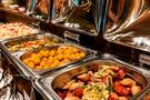 Teplý bufet s nerezovými ohřívacími pánvemi: klobásky s cibulí, zlaté bramborové krokety a pečená zelenina.