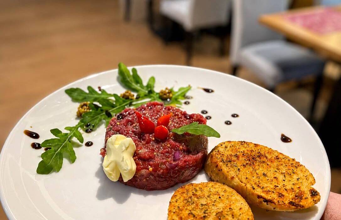 Hovězí tatarák s rukolou, hořčičnými perličkami, krémem a dvěma bylinkovými crostini na talíři v restauraci