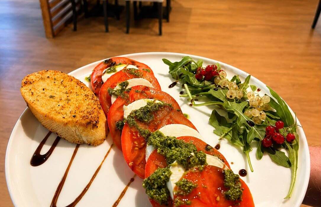 Caprese s rajčetem, mozzarellou, pestem, balsamikem, rukolou a česnekovým toustem na talíři v hotelové restauraci.