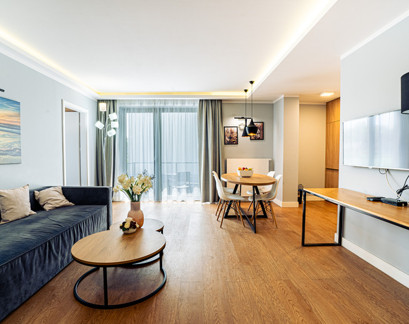Moderní obývací prostor v apartmánu se semišovou pohovkou, kulatými dřevěnými stolky, jídelním stolem a balkonem za záclonami