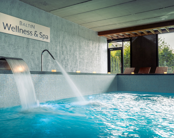 Vnitřní bazén Baltin Wellness & Spa s vodopádovými tryskami, mozaikou, dřevěnými lehátky, svíčkami a výhledem do zahrady