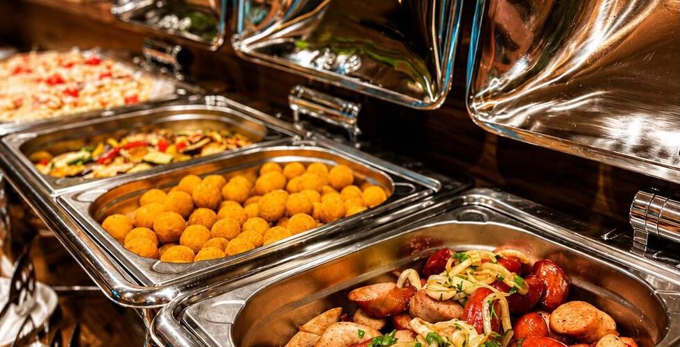 Teplý bufet s nerezovými ohřívacími pánvemi: klobásky s cibulí, zlaté bramborové krokety a pečená zelenina.