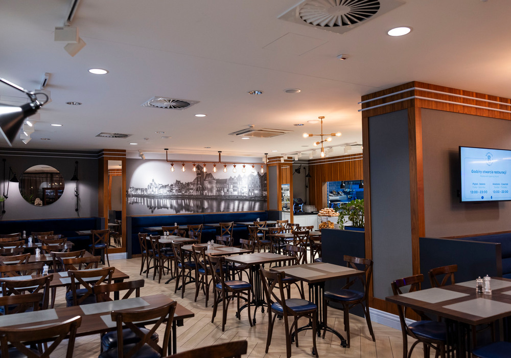 Hotel dining room with wooden tables, navy banquette seating, pendant lights and black‑and‑white waterfront mural.