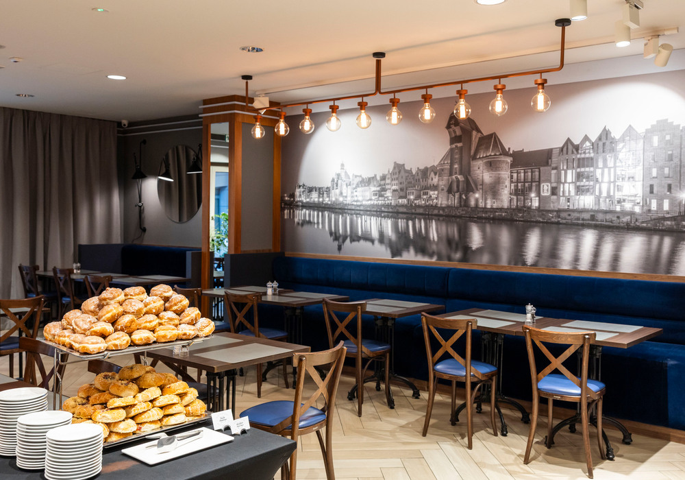 Breakfast buffet with stacked pastries and plates in a modern hotel dining room with navy banquette, wooden chairs and waterfront mural