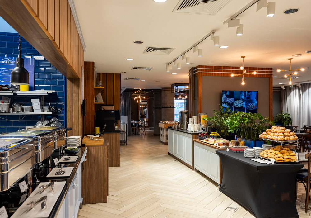 Breakfast buffet with chafing dishes, stacked pastries, cereal dispensers and coffee in a bright dining area with wood accents.