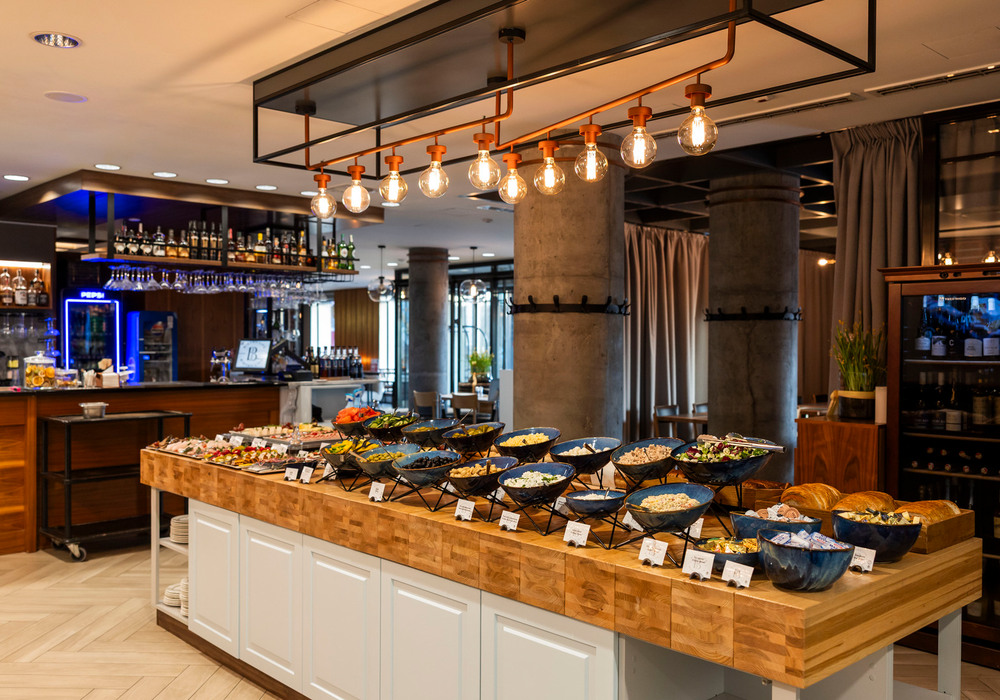 Hotel buffet spread on wooden island with bowls of salads, breads and condiments, warm pendant lights and a bar in background