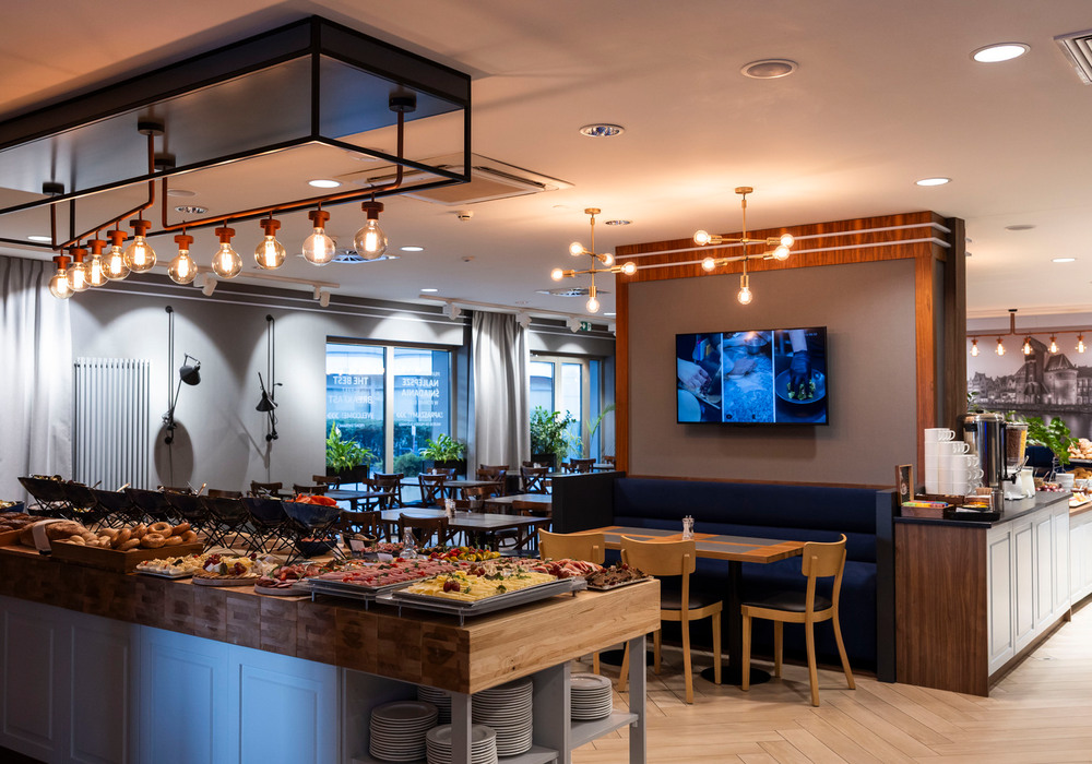 Breakfast buffet with breads, cheeses and cold cuts on a wooden island, coffee station, booth seating and warm pendant lights