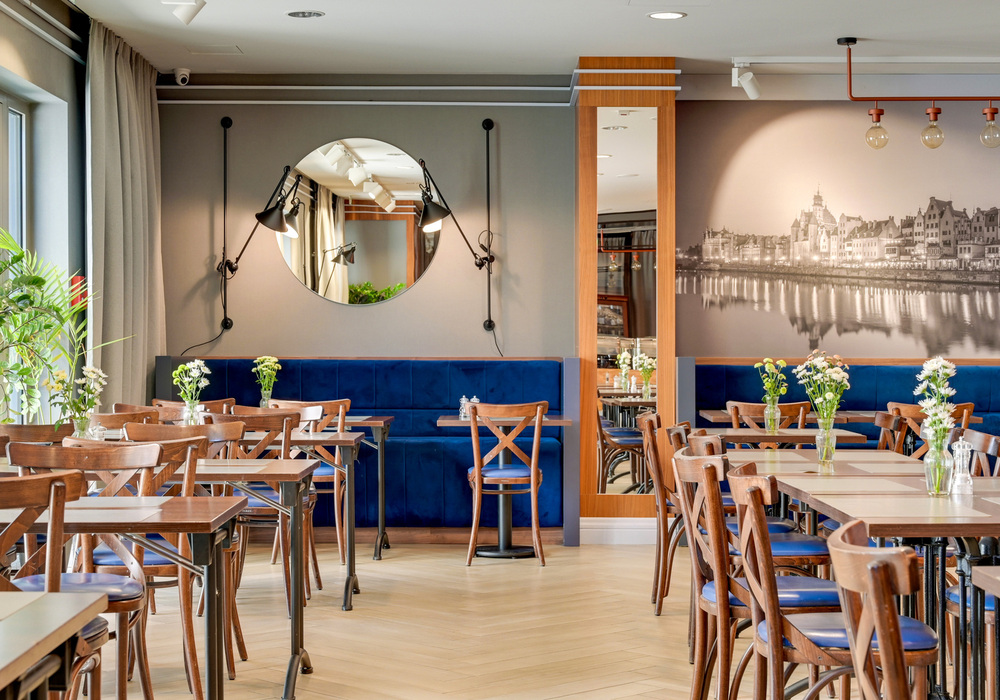 Bright hotel dining room with blue velvet banquette, wooden tables and chairs, round wall mirror, cityscape mural, fresh flowers and natural light