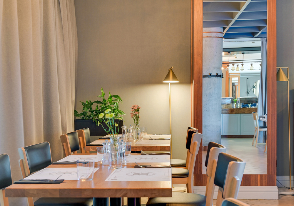 Contemporary hotel dining room with wooden tables set for guests, soft floor lamp, potted plants and mirrored view of buffet.
