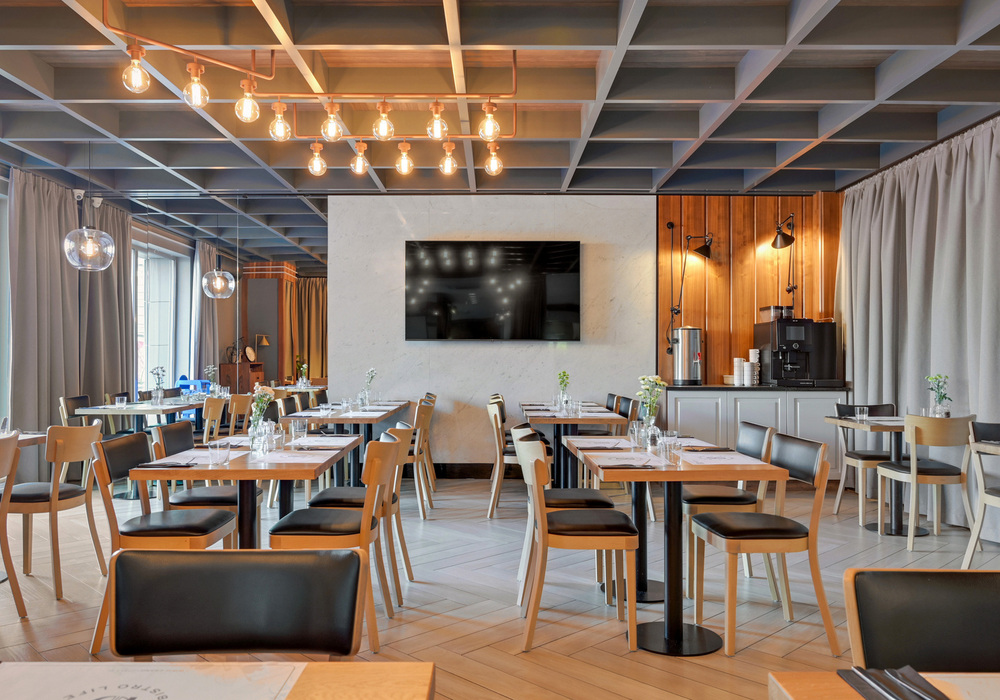 Contemporary hotel dining room with wooden tables set for meals, black leather chairs, hanging pendant lights, TV and coffee station.