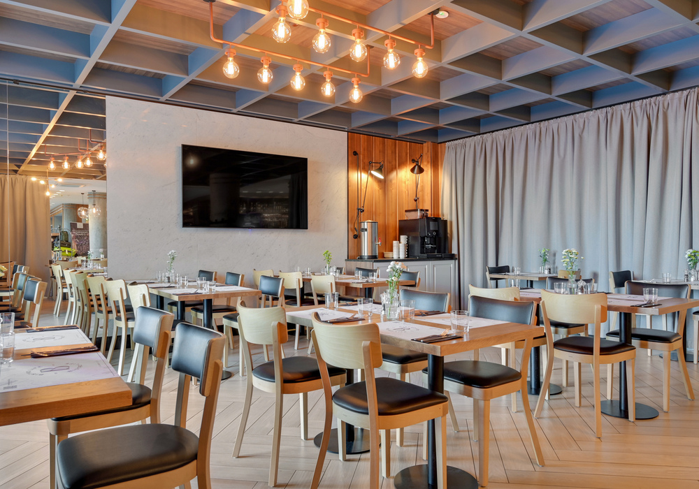 Modern hotel restaurant with wooden tables and chairs set for dining, filament pendant lights, wall TV and coffee station