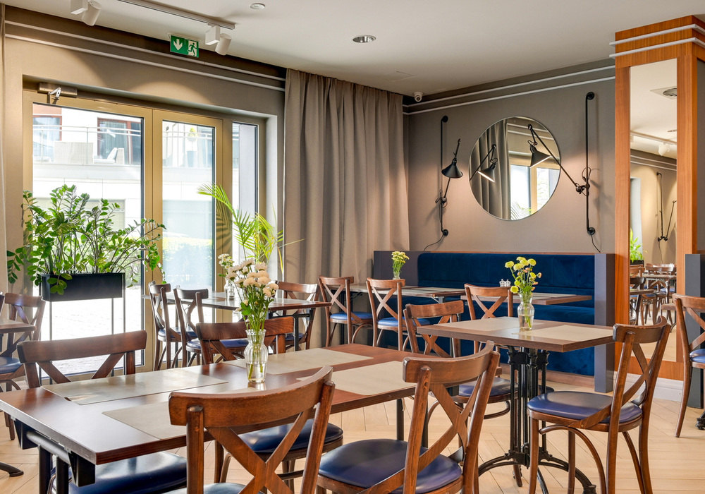 Hotel dining area with wooden tables and chairs, blue banquette seating, vases of fresh flowers, large windows and indoor plants