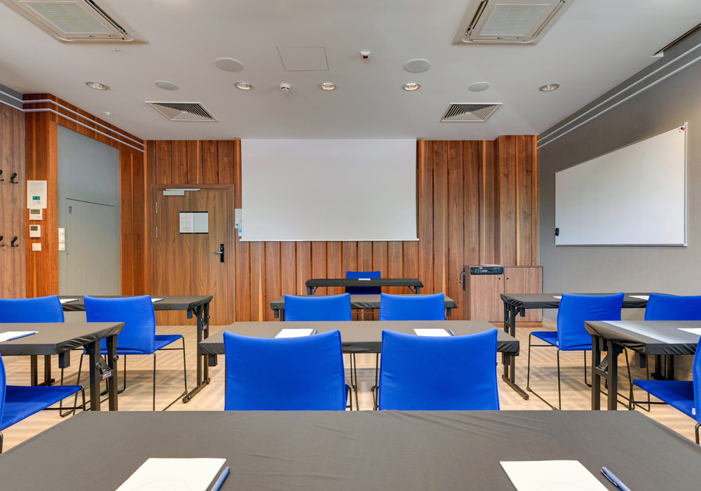 Hotel meeting room with classroom-style tables, bright blue chairs, wooden paneled wall, projector screen and whiteboard
