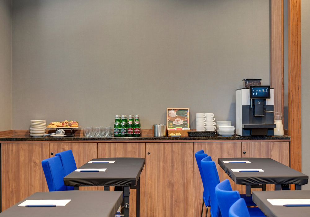Conference refreshment counter with coffee machine, milk jug, stacked cups, bottled water, pastries and meeting tables with notepads.