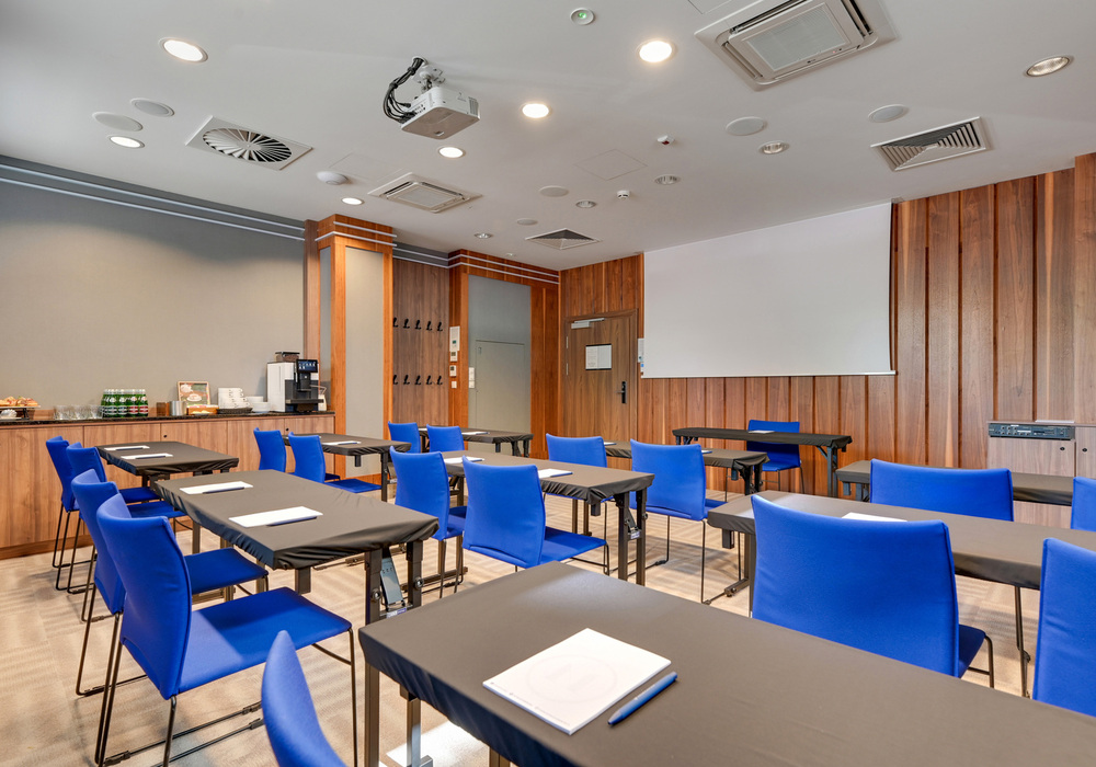 Conference room with blue chairs, classroom tables, ceiling projector and screen, wooden walls and coffee station.