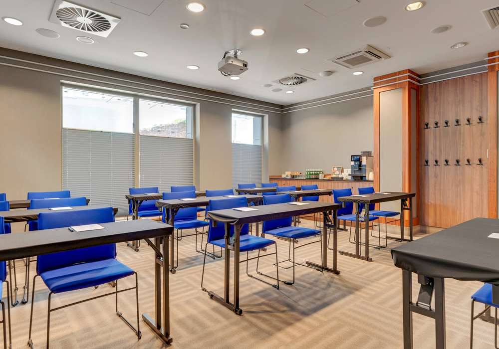 Conference room with rows of tables and blue chairs, ceiling projector and a coffee/snack station at the back