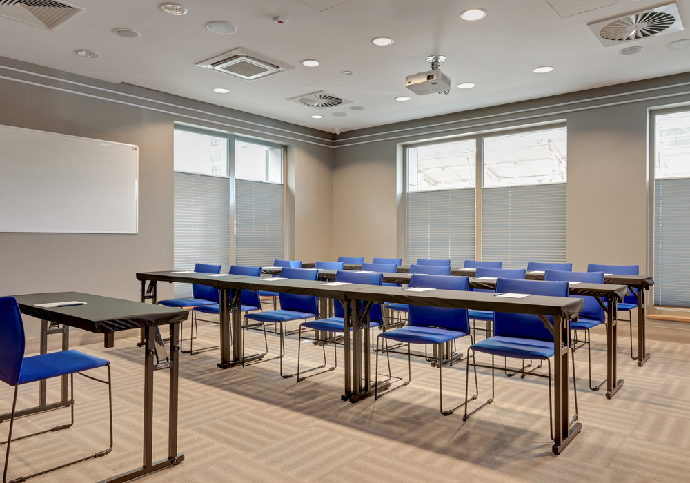 Bright hotel meeting room with rows of black tables, blue chairs, ceiling projector and whiteboard