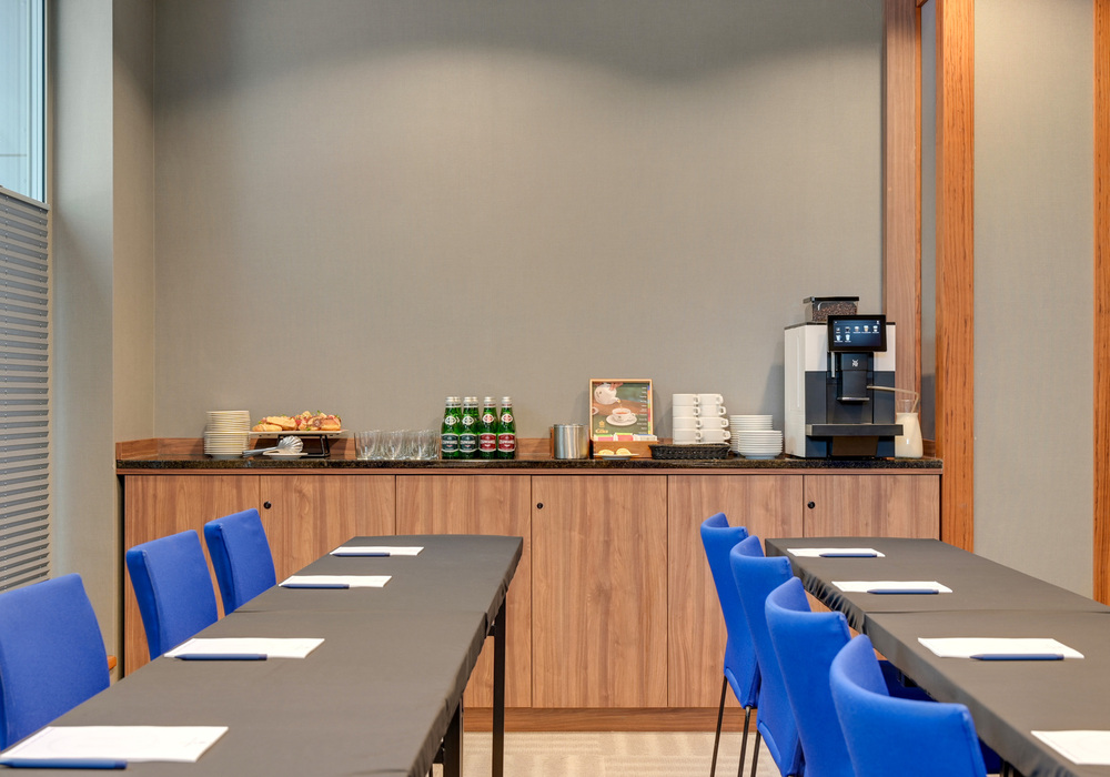 Conference room buffet on a wooden counter: espresso machine, bottled water, pastries and cups; tables with black covers and blue chairs.