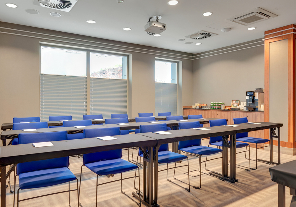 Classroom-style hotel meeting room with blue chairs, long tables, ceiling projector and coffee/snack station