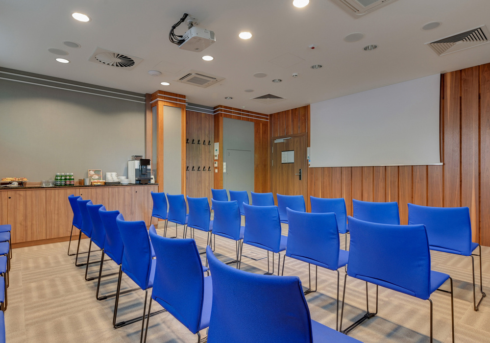 Small hotel conference room with blue theater chairs, projector and screen, wood-paneled walls and coffee station
