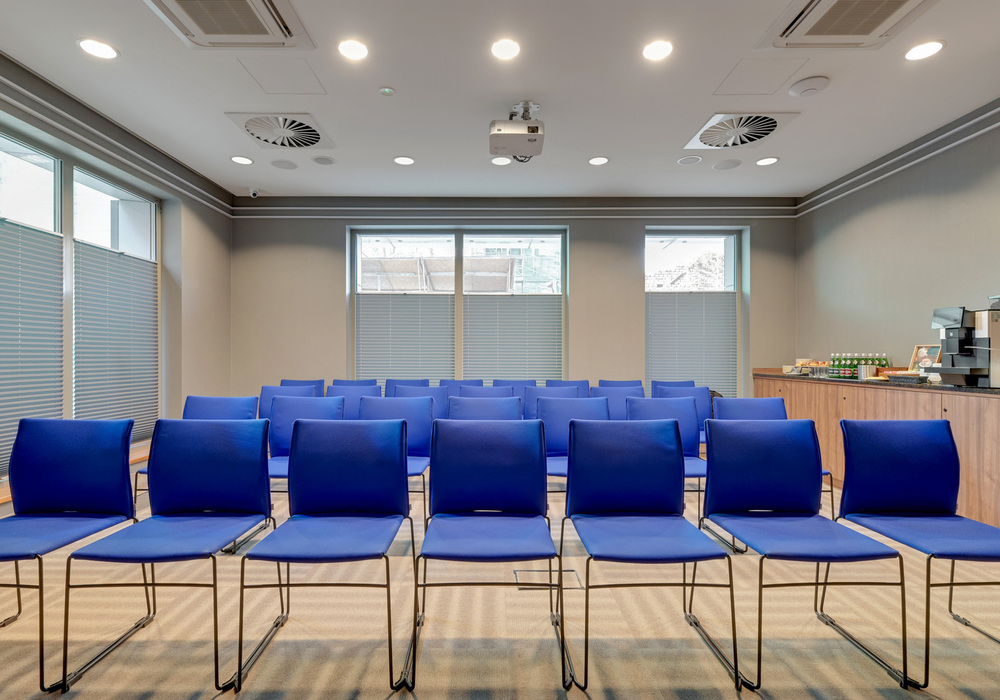 Conference room with rows of blue chairs, ceiling projector, coffee and snack station, window blinds and neutral modern decor