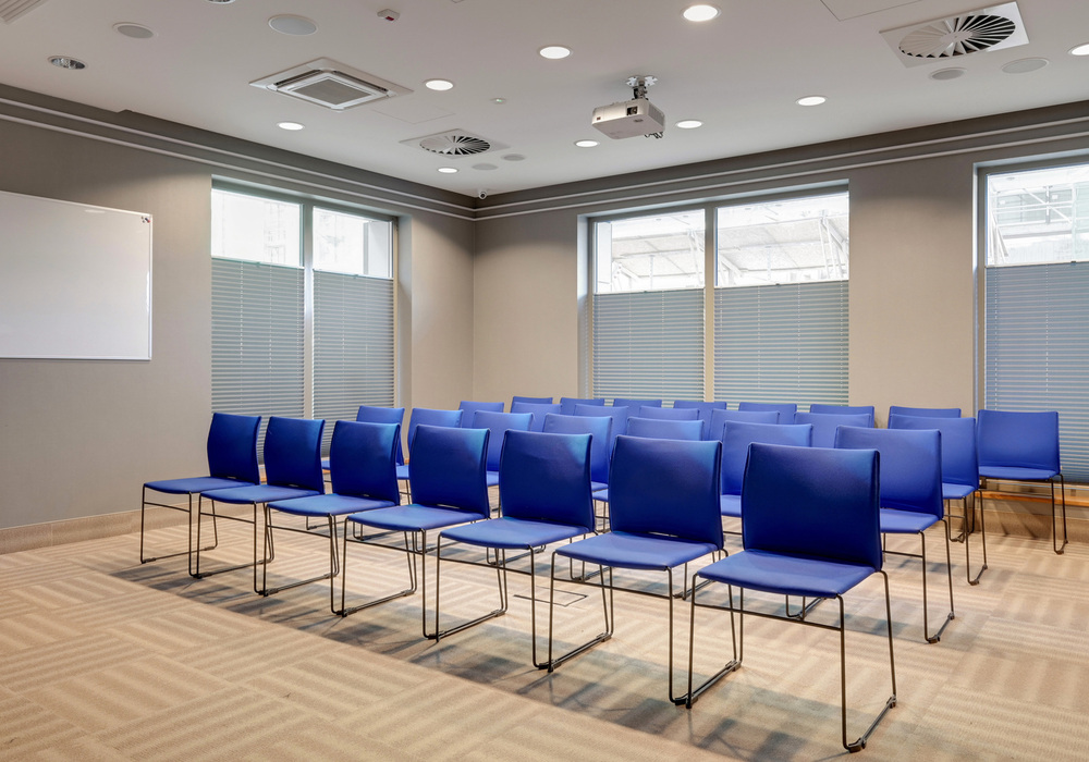 Conference room with rows of blue chairs, ceiling projector, whiteboard, blinds and neutral décor for meetings and training