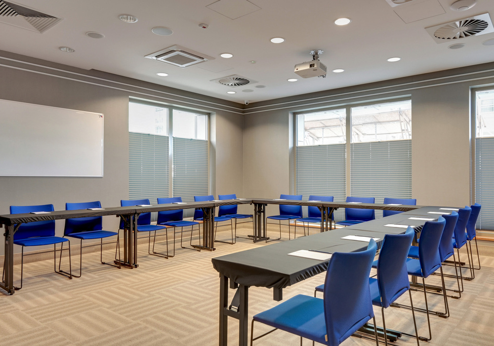 Bright modern meeting room with U-shaped tables, blue chairs, ceiling projector and whiteboard, soft natural light.