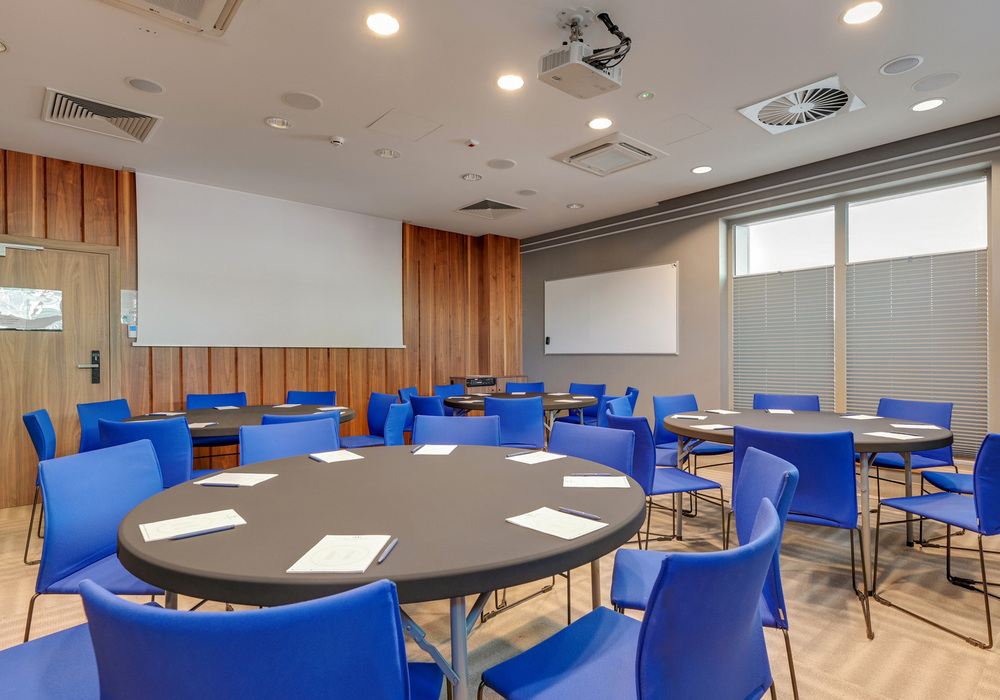 Conference room with round tables, blue chairs, ceiling projector, whiteboard and wood-paneled wall