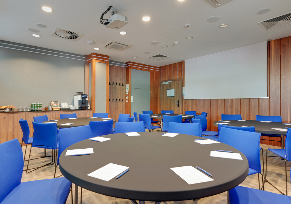 Hotel meeting room with round tables, blue chairs, notepads and pens, projector, screen and coffee station