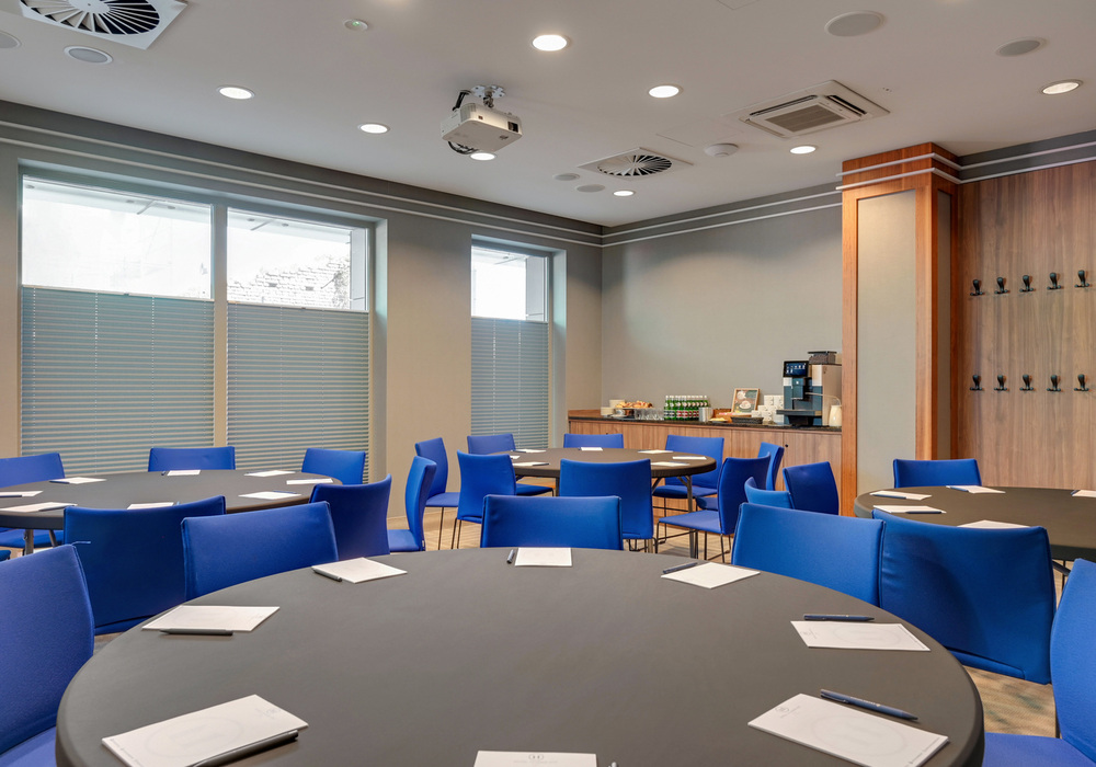 Conference room with round tables, bright blue chairs, ceiling projector, notepads and pens, and a coffee/refreshment station.